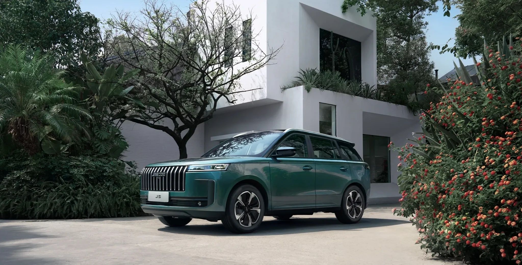Jaecoo J5 SUV parked outside a modern luxury home with lush landscaping.