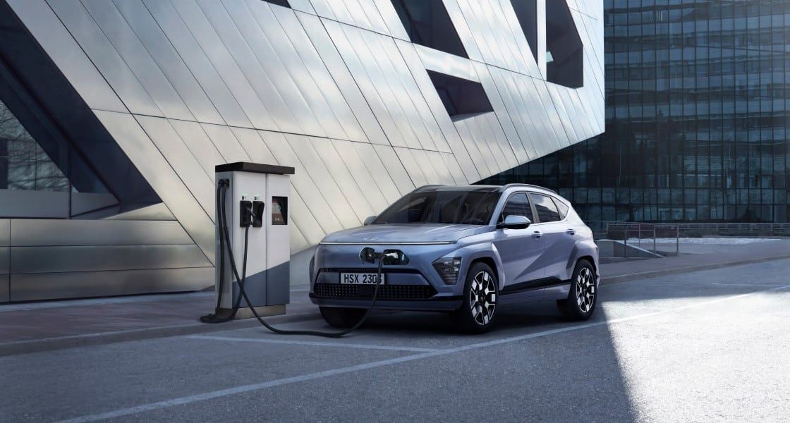 Hyundai Kona Electric charging at a public EV charging station outside a modern commercial building.