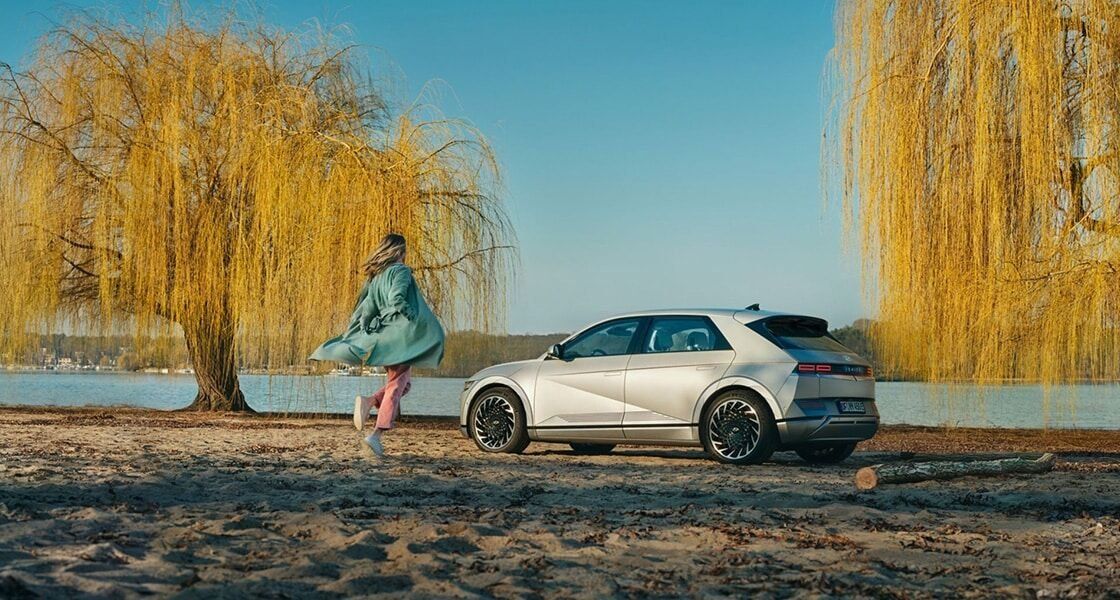 Hyundai IONIQ 5 parked beside a lakeside with willow trees and a woman walking nearby.